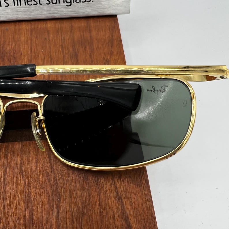 Puede incluir: Gafas de sol con montura dorada, patillas negras y lentes verde oscuro. Las gafas de sol est&aacute;n sobre una superficie de madera. Las lentes tienen una ligera curvatura y la montura es rectangular.