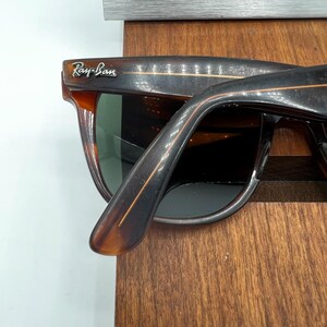 May include: Close-up of Ray-Ban sunglasses with a brown tortoiseshell frame. The sunglasses have dark lenses and the Ray-Ban logo is visible on the upper left corner. The frame is resting on a wooden surface.