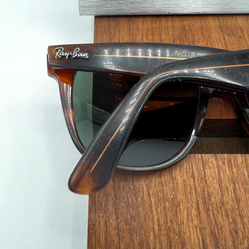 May include: Close-up of Ray-Ban sunglasses with a brown tortoiseshell frame. The sunglasses have dark lenses and the Ray-Ban logo is visible on the upper left corner. The frame is resting on a wooden surface.