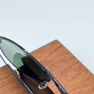 Pu&ograve; includere: Occhiali da sole neri con lenti verdi. Gli occhiali da sole sono su una superficie di legno. Il testo "MADE IN ITALY" &egrave; visibile sulla montatura.