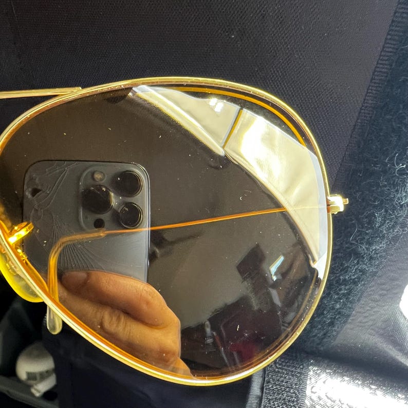 Puede incluir: Gafas de sol tipo aviador con una montura fina de metal dorado y lentes tintadas en marr&oacute;n oscuro. La superficie reflectante de la lente muestra el reflejo n&iacute;tido de un smartphone con sistema de triple c&aacute;mara y parte de una mano sosteniendo el dispositivo.