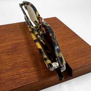 May include: Foldable sunglasses with a tortoiseshell pattern in shades of brown, black, and yellow. The lenses are a light brown color. The sunglasses are resting on a wooden surface.
