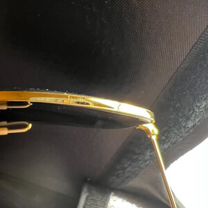 May include: Close-up of gold-rimmed sunglasses with dark lenses. The gold frame has a shiny finish, and the lenses are a deep black. The sunglasses are set against a dark, textured background.