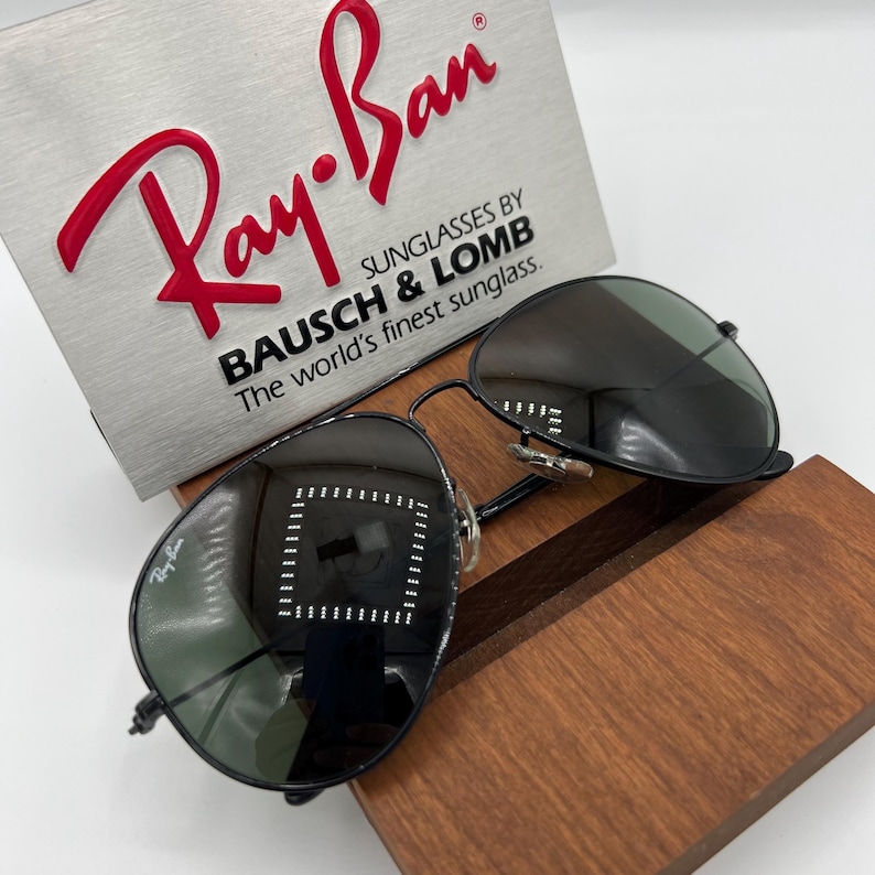 Pu&ograve; includere: Occhiali da sole Ray-Ban neri con un logo Ray-Ban argentato sulla custodia. Gli occhiali da sole sono su un espositore in legno. La custodia riporta la scritta "Sunglasses by Bausch & Lomb. The world's finest sunglasses."