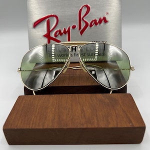 K&ouml;nnte beinhalten: Goldene Aviator-Sonnenbrille mit gr&uuml;nen Gl&auml;sern. Die Sonnenbrille liegt auf einem Holzst&auml;nder. Das Ray-Ban-Logo ist auf einer silbernen Box hinter der Sonnenbrille zu sehen.