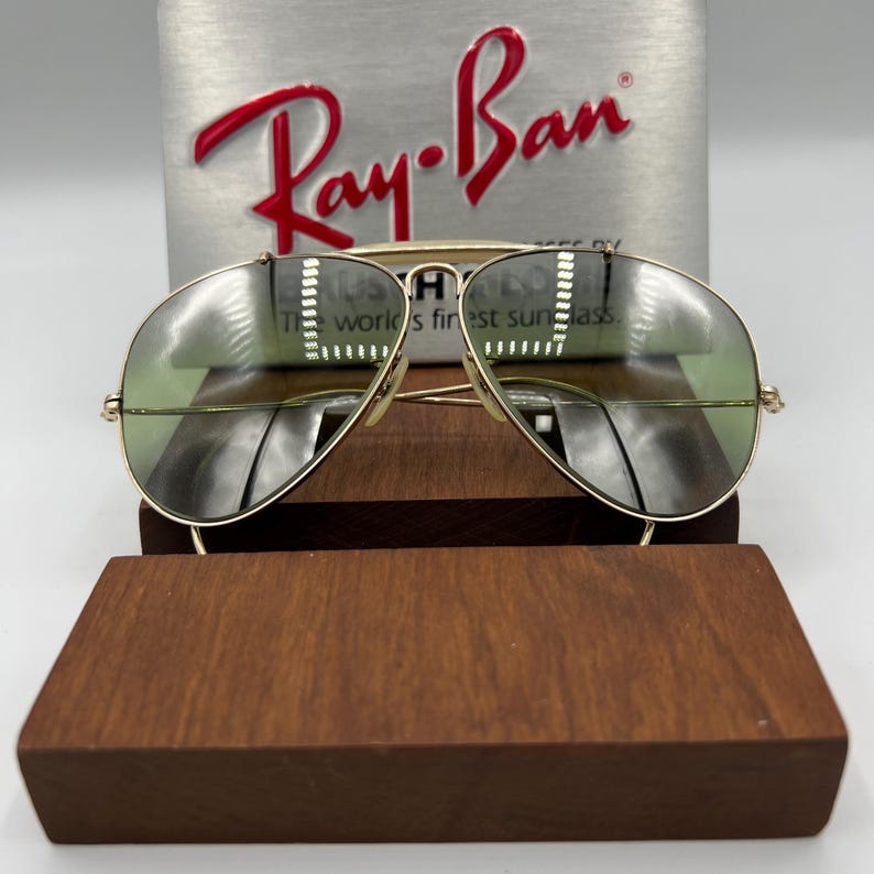 K&ouml;nnte beinhalten: Goldene Aviator-Sonnenbrille mit gr&uuml;nen Gl&auml;sern. Die Sonnenbrille liegt auf einem Holzst&auml;nder. Das Ray-Ban-Logo ist auf einer silbernen Box hinter der Sonnenbrille zu sehen.