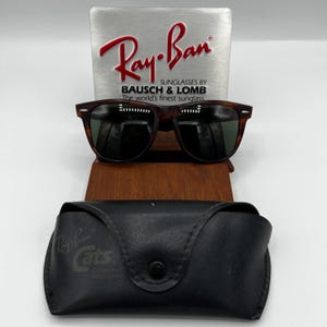 May include: Brown tortoiseshell sunglasses with dark lenses, displayed on a wooden stand. The Ray-Ban logo is visible on a silver sign behind the glasses. A black leather case with the Ray-Ban Cats logo is in front.