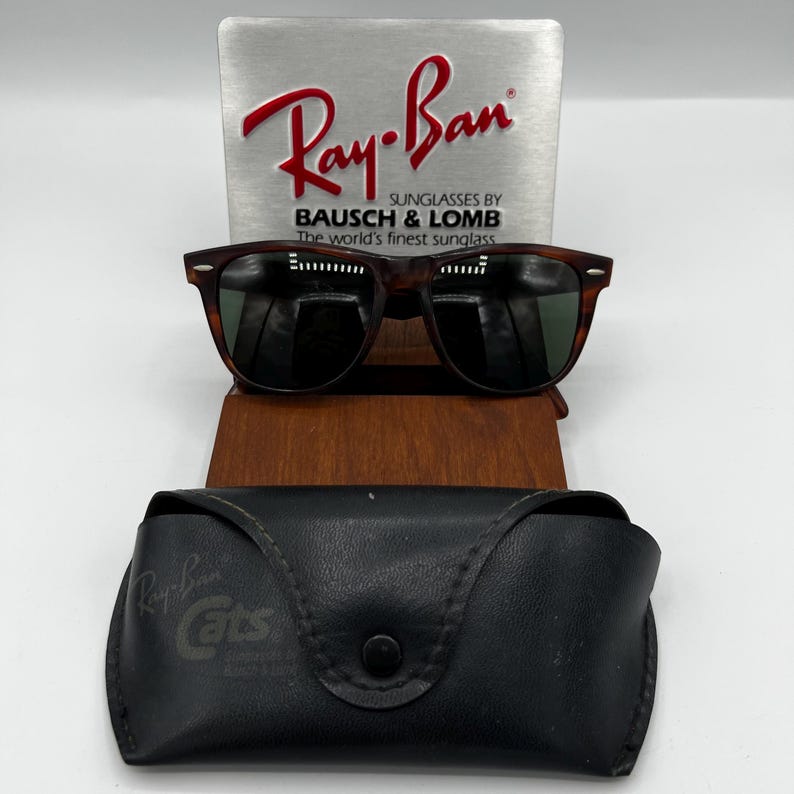 May include: Brown tortoiseshell sunglasses with dark lenses, displayed on a wooden stand. The Ray-Ban logo is visible on a silver sign behind the glasses. A black leather case with the Ray-Ban Cats logo is in front.
