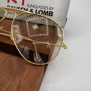 May include: Gold aviator sunglasses with brown lenses. The sunglasses are resting on a wooden surface. The brand name "BAUSCH & LOMB" is visible on a silver label behind the sunglasses.