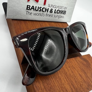 Puede incluir: Gafas de sol marrones oscuras con un patr&oacute;n de carey. Las gafas de sol se exhiben en un soporte de madera. El texto en el letrero dice: "SUNGLASSES BY BAUSCH & LOMB The world's finest sunglass."