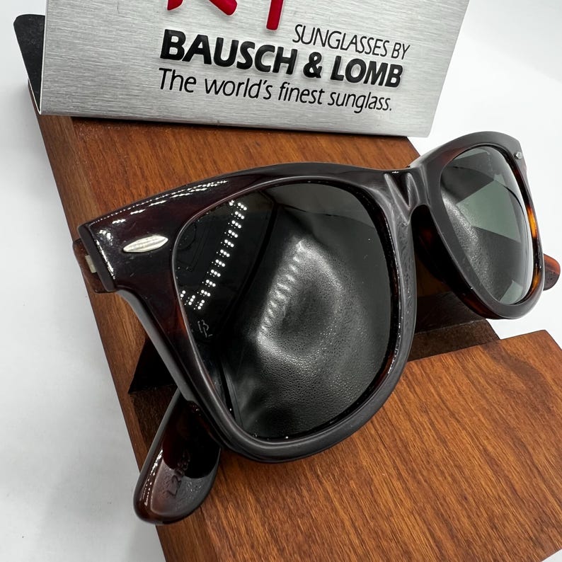 Puede incluir: Gafas de sol marrones oscuras con un patr&oacute;n de carey. Las gafas de sol se exhiben en un soporte de madera. El texto en el letrero dice: "SUNGLASSES BY BAUSCH & LOMB The world's finest sunglass."