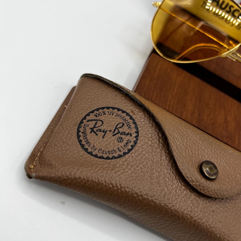 Puede incluir: Un estuche para gafas de cuero texturizado color canela con un sello circular negro de Ray-Ban que dice 100% UV protection Sunglasses by Bausch & Lomb. Unas gafas de sol con montura dorada y lentes &aacute;mbar descansan sobre un soporte de madera oscura.