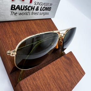 May include: Gold-framed sunglasses with dark lenses rest on a wooden display stand. The sunglasses have a classic aviator style. The text "SUNGLASSES BY BAUSCH & LOMB The world's finest sunglass" is visible above the sunglasses.