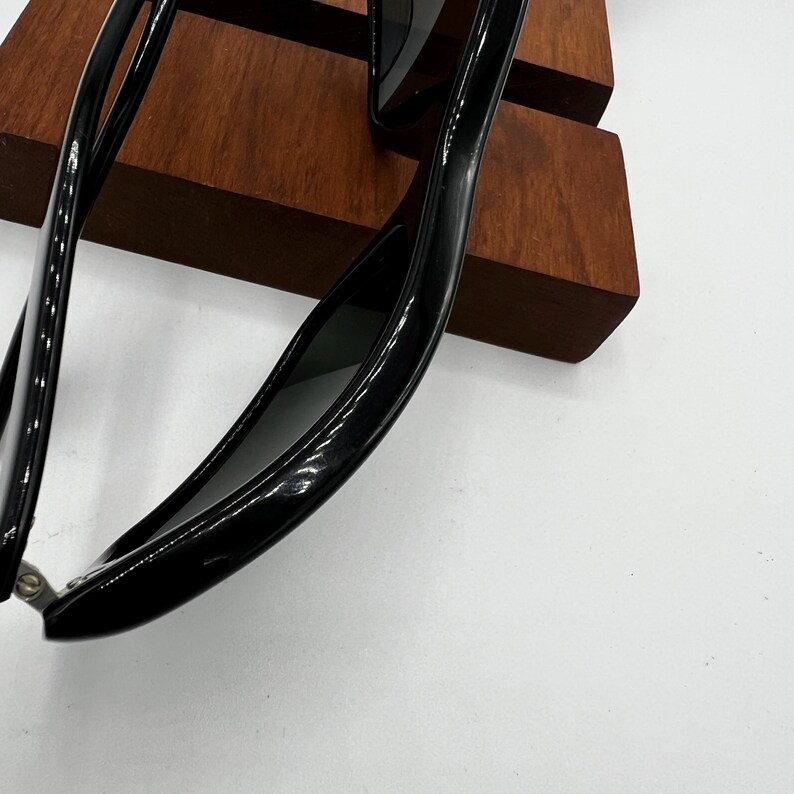 May include: Close-up of black sunglasses with a glossy finish. The sunglasses are resting on a wooden display stand. The lenses are dark, and the frames have a sleek, modern design. The background is white.