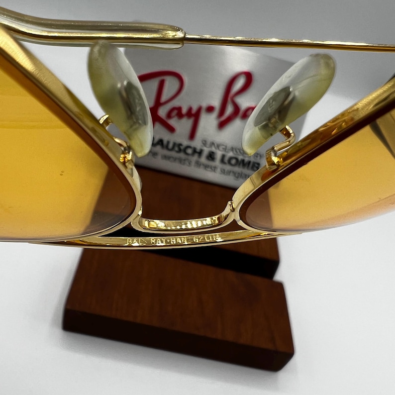 Puede incluir: Gafas de sol de metal dorado con lentes tintadas en &aacute;mbar y almohadillas nasales transparentes. El puente est&aacute; grabado con B&L RAY-BAN 62[]14. Al fondo, un cartel blanco muestra el logo rojo de Ray-Ban y el texto BAUSCH & LOMB the world's finest sunglasses.