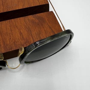 Pu&ograve; includere: Occhiali da sole con montatura marmorizzata verde scuro e nera e lenti scure. Gli occhiali da sole hanno aste e cerniere in metallo color oro. Gli occhiali da sole sono appoggiati su una superficie di legno.