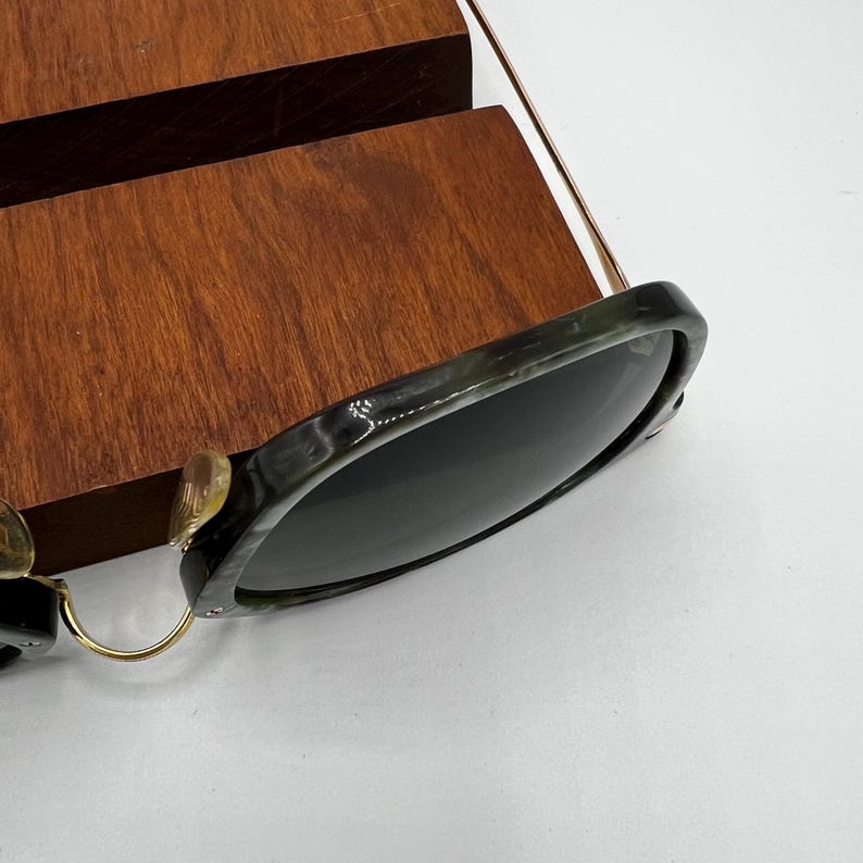 Pu&ograve; includere: Occhiali da sole con montatura marmorizzata verde scuro e nera e lenti scure. Gli occhiali da sole hanno aste e cerniere in metallo color oro. Gli occhiali da sole sono appoggiati su una superficie di legno.