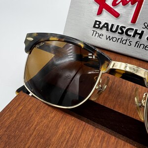 May include: Tortoiseshell-framed sunglasses with brown lenses. The sunglasses have gold-colored metal accents and the Ray-Ban logo is visible on the frame. The text "BAUSCH" and "The world's finest" are also visible.