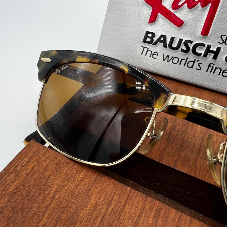 May include: Tortoiseshell-framed sunglasses with brown lenses. The sunglasses have gold-colored metal accents and the Ray-Ban logo is visible on the frame. The text "BAUSCH" and "The world's finest" are also visible.