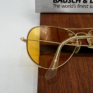 Puede incluir: Gafas de sol tipo aviador en tono dorado con lentes tintadas en &aacute;mbar y puntas de patillas transparentes. La montura incluye un dise&ntilde;o de doble puente. Un letrero parcial al fondo muestra el texto Bausch & Lomb y The world's finest sunglasses.