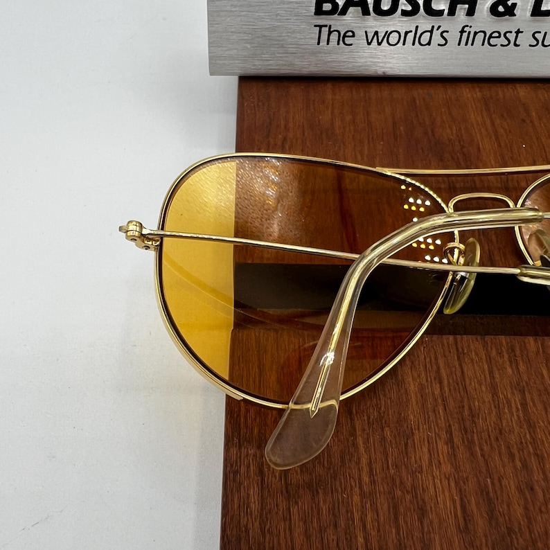 Puede incluir: Gafas de sol tipo aviador en tono dorado con lentes tintadas en &aacute;mbar y puntas de patillas transparentes. La montura incluye un dise&ntilde;o de doble puente. Un letrero parcial al fondo muestra el texto Bausch & Lomb y The world's finest sunglasses.