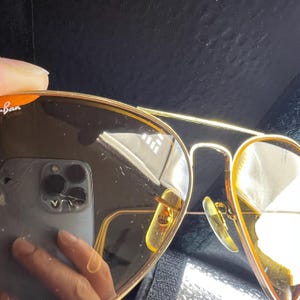 Puede incluir: Gafas de sol tipo aviador de metal en tono dorado con lentes tintadas en marr&oacute;n. La lente izquierda muestra el logotipo blanco de Ray-Ban en la esquina superior. La montura incluye un dise&ntilde;o de doble puente y almohadillas nasales ajustables.