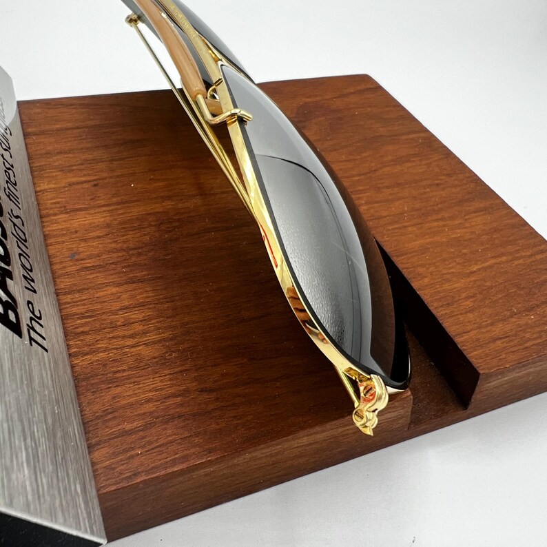 May include: Sunglasses with a gold frame and dark lenses, resting on a wooden display stand. The sunglasses have a sleek, modern design with a gold bridge and temples. The lenses are a dark, reflective color. The text "The world's finest sung" is partially visible.