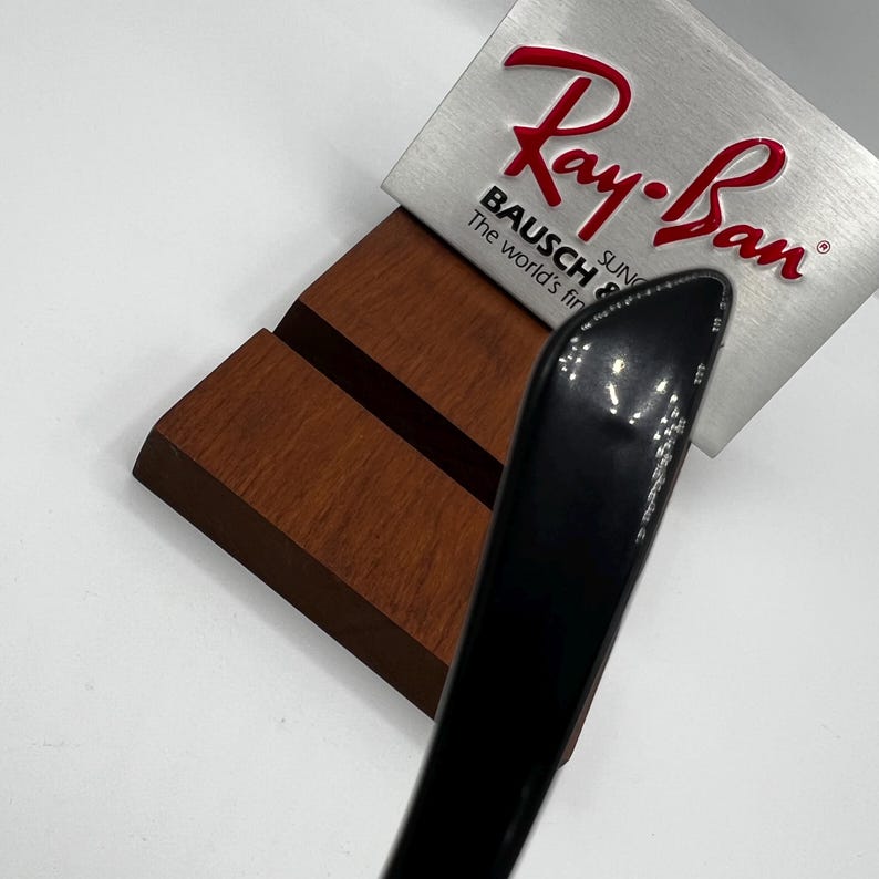 May include: A black sunglasses arm rests on a wooden display stand. A silver sign with the red "Ray-Ban" logo and the text "BAUSCH & LOMB SUN The world's finest" is behind the sunglasses.