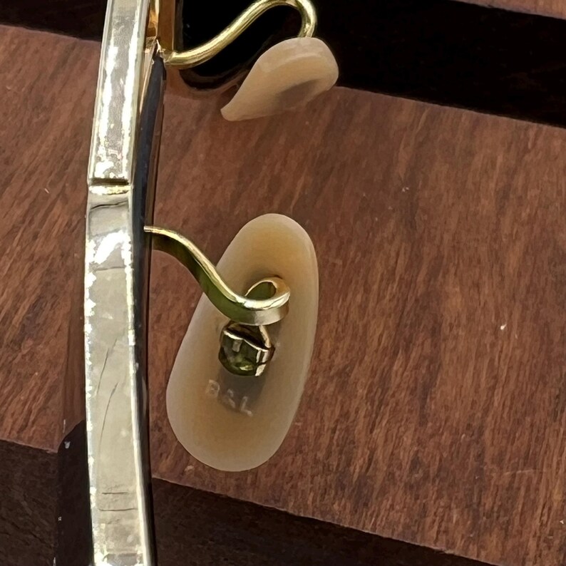 K&ouml;nnte beinhalten: Nahaufnahme der goldfarbenen Metallb&uuml;gelspitze einer Brille. Die Spitze ist an einem goldfarbenen Metallb&uuml;gel befestigt. Die Spitze hat eine transparente Kunststoffabdeckung mit dem aufgedruckten Wort "BAL".