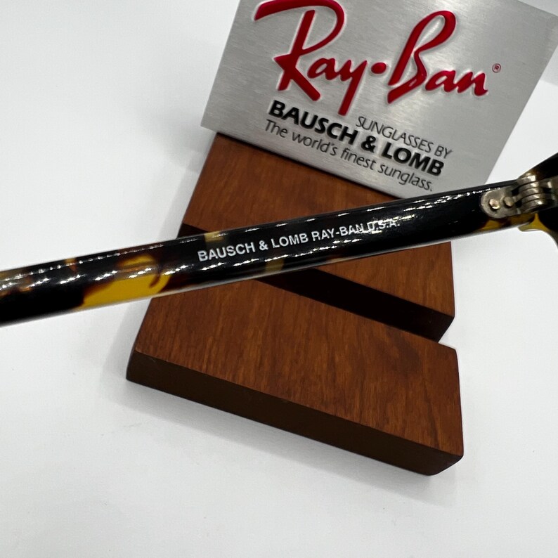 May include: Close-up of Ray-Ban sunglasses with a tortoiseshell pattern on the arm. The text "Ray-Ban" is displayed in red, and the brand name "BAUSCH & LOMB" is also visible. The sunglasses are resting on a wooden display stand.