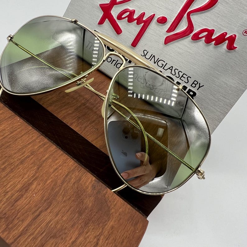 K&ouml;nnte beinhalten: Goldene Aviator-Sonnenbrille mit gr&uuml;nen Gl&auml;sern. Die Sonnenbrille liegt auf einer Holzoberfl&auml;che. Das Ray-Ban-Logo ist auf der Schachtel im Hintergrund sichtbar. Sonnenbrillen von Ray-Ban.