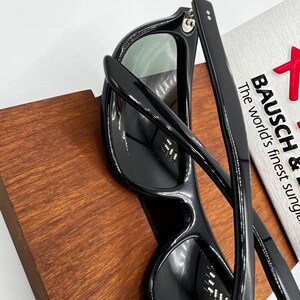 May include: Black sunglasses with a rectangular frame and green lenses. The sunglasses are resting on a wooden surface. The text "BAUSCH &" and "The world's finest sung" is visible on a silver card.