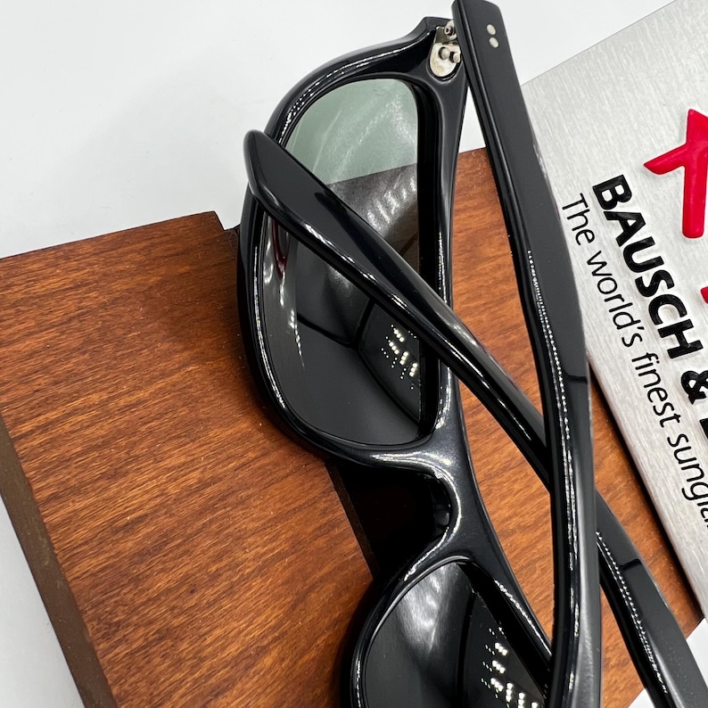 May include: Black sunglasses with a rectangular frame and green lenses. The sunglasses are resting on a wooden surface. The text "BAUSCH &" and "The world's finest sung" is visible on a silver card.