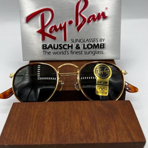 May include: Gold-rimmed Ray-Ban sunglasses with dark lenses, resting on a wooden display. The Ray-Ban logo is visible on the top left lens. The sunglasses have a classic round shape with tortoise-shell arms. The brand name and "Sunglasses by Bausch & Lomb" are displayed above.