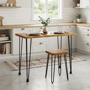 Puede incluir: Una mesa rectangular de madera y un taburete cuadrado a juego, ambos con patas de horquilla de metal negro. La superficie de la mesa es de madera marrón claro, con dos platos y dos vasos. El taburete tiene un asiento de madera a juego.