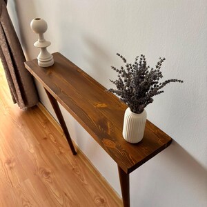 May include: A long, narrow wooden console table with a dark brown stained finish and tapered legs. On the surface sits a white, ribbed ceramic vase filled with dried lavender sprigs and a separate white, sculptural, multi-tiered candle holder.