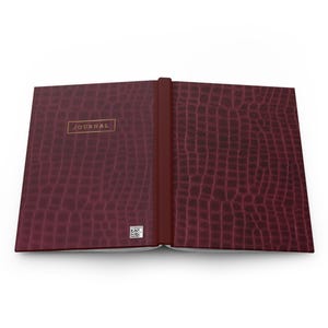 May include: A dark burgundy journal with a textured crocodile skin pattern. The front cover features a rectangular gold-outlined label with the word JOURNAL printed in gold. A small square QR code is visible near the bottom of the front cover.