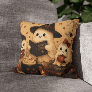 May include: A square throw pillow featuring a beige background with a cartoon illustration of three ghosts. One ghost wears a witch hat and reads a book titled SPOOKY STORIES. Another holds a pumpkin, and a third holds a mug with whipped cream.