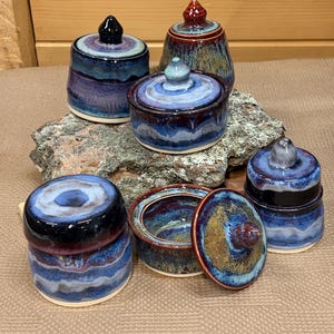 May include: A collection of six ceramic lidded jars and bowls featuring a glossy glaze in shades of deep blue, purple, and earthy brown. Each piece has a distinct, hand-thrown appearance with smooth, rounded lids and a consistent color palette.