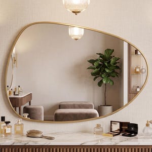 May include: An asymmetrical wall mirror with a thin gold frame mounted above a marble-topped vanity. The mirror reflects a room with a potted plant, a stool, and a dressing table. Two gold cylindrical wall sconces flank the mirror on the wall.