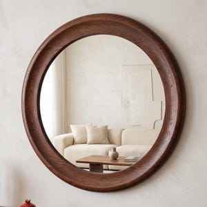 May include: A large circular wall mirror featuring a thick, dark brown wooden frame with a visible natural wood grain texture. The mirror reflects a room interior containing a beige sofa, a wooden coffee table, and a small ceramic vase.