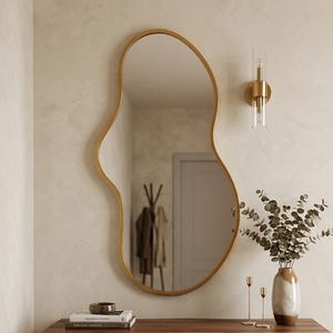 May include: An irregular, organic-shaped wall mirror with a thin gold frame. Below the mirror sits a rectangular console table featuring a natural wood grain top and four slender gold hairpin legs, holding a ceramic vase with eucalyptus branches.
