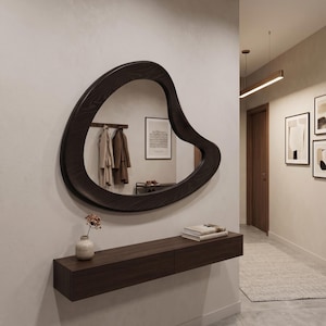 May include: An irregular, organic-shaped wall mirror with a dark wood frame featuring a visible grain pattern. The mirror is mounted above a rectangular, dark wood floating console shelf that holds a small ceramic vase and a stack of books.