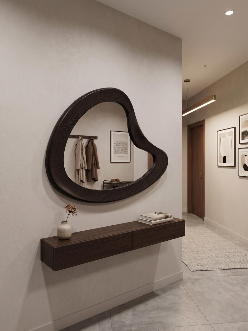 May include: An irregular, organic-shaped wall mirror with a dark wood frame featuring a visible grain pattern. The mirror is mounted above a rectangular, dark wood floating console shelf that holds a small ceramic vase and a stack of books.
