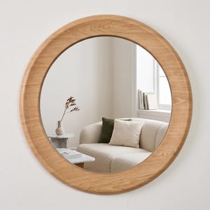 May include: A circular wall mirror featuring a wide, natural light-toned wooden frame with a visible grain pattern. The mirror surface reflects a room interior containing a beige sofa with cushions, a small side table, and a vase with a plant.