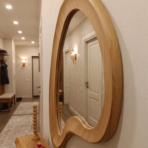 May include: A wall-mounted mirror featuring an irregular, organic-shaped frame crafted from light-toned wood with a visible grain pattern. The mirror is positioned on a white wall above a wooden console table, reflecting a hallway interior.