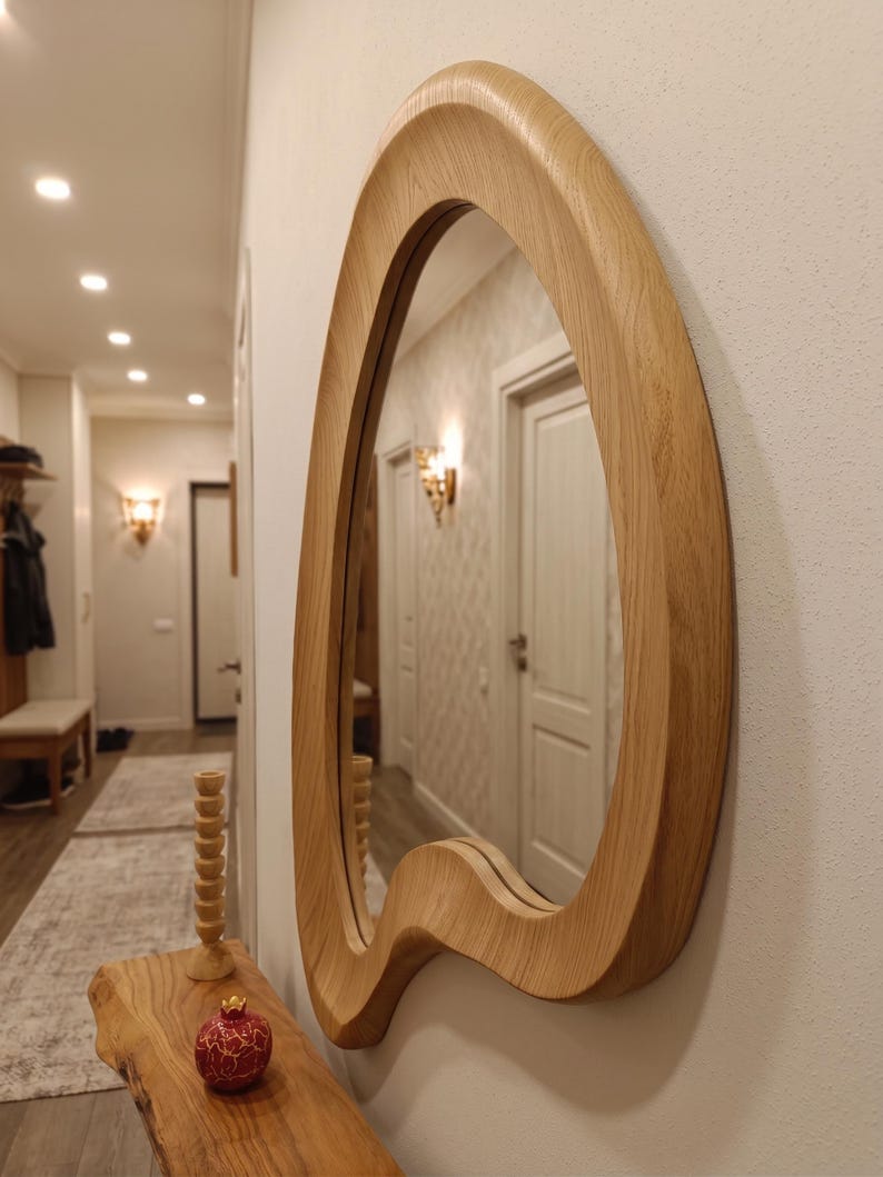 May include: A wall-mounted mirror featuring an irregular, organic-shaped frame crafted from light-toned wood with a visible grain pattern. The mirror is positioned on a white wall above a wooden console table, reflecting a hallway interior.