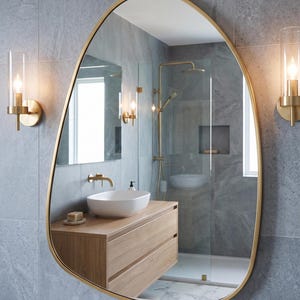 May include: An asymmetrical wall mirror with a thin gold-toned metal frame. The mirror has an irregular, organic shape and is mounted on a gray tiled wall between two cylindrical glass wall sconces with gold-toned hardware and warm light.