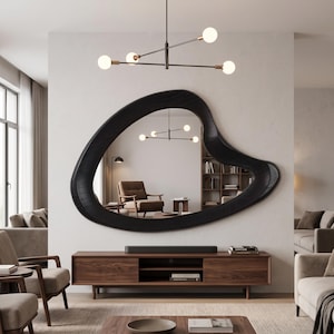 May include: A large, black, organic-shaped wall mirror with a thick, textured frame. The mirror reflects a living room interior, including a wooden media console, a brown armchair, a bookshelf, and a modern four-light chandelier hanging above.