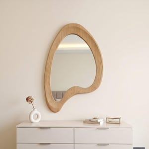May include: A wall-mounted mirror with an irregular, organic, amoeba-like shape and a light-toned wood grain frame. The mirror features a smooth, curved wooden border that surrounds the reflective glass surface, creating a distinct, asymmetrical silhouette.